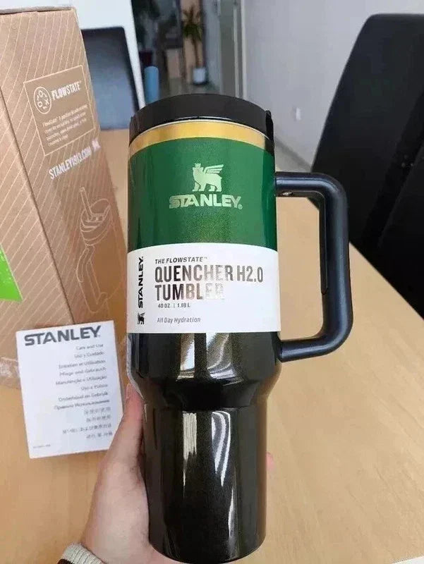 Black Stanley tumbler with green lid held by a hand on a wooden surface.