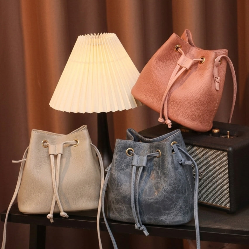 Three drawstring bags in beige, blue, and pink on a dark surface with a lamp and speaker in the background.